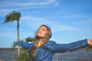 Blonde woman wearing a jean jacket and has reef safe sunscreen on her nose. She is  spreading her arms and looking toward the sky. The background is a blue sky with palm trees. The value proposition is written in the background.